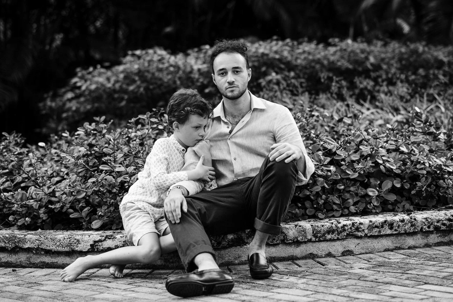 A young man with short dark hair, wearing a light-colored shirt and dark pants, sitting on a stone ledge next to a young girl with short dark hair, a patterned shirt, and shorts, who is leaning on him with her head resting on his shoulder in a park w