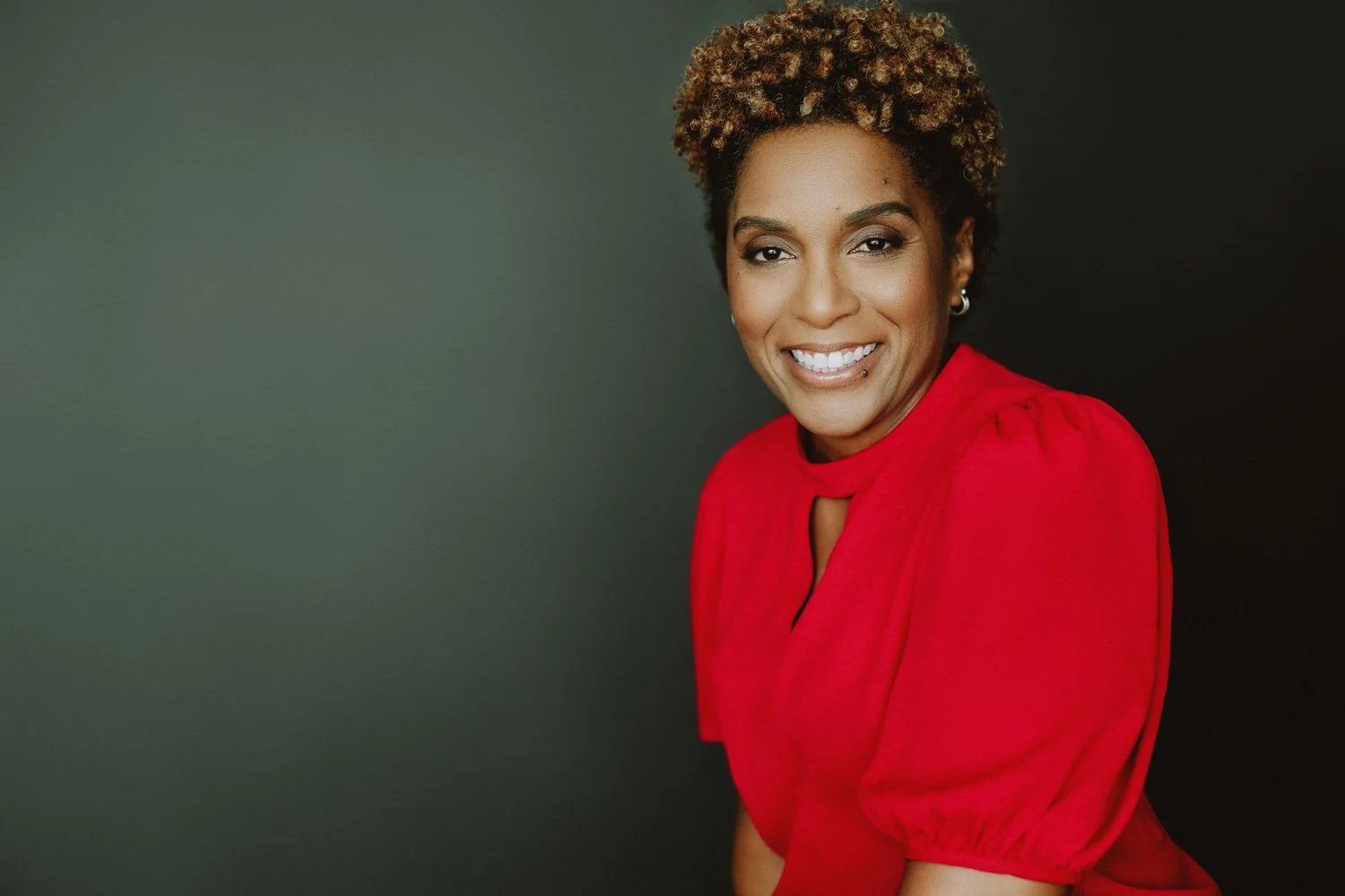 A smiling woman with short curly hair, wearing a red dress, posing against a dark background.