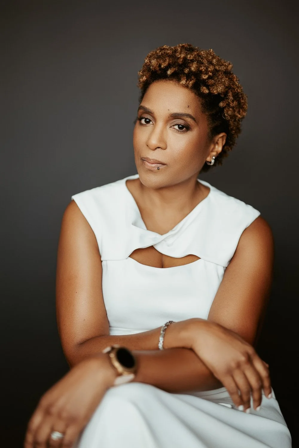 A woman with short, curly brown hair, wearing a white sleeveless dress with a unique neckline, sitting against a dark gray background. She has jewelry including earrings, a bracelet, and a watch, and shows a calm, confident expression.