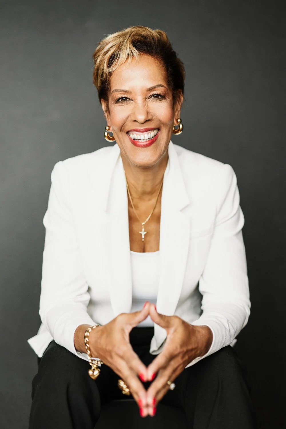 An older woman smiling warmly, wearing a white blazer and black pants, with gold jewelry and a cross necklace, sitting against a dark gray background.