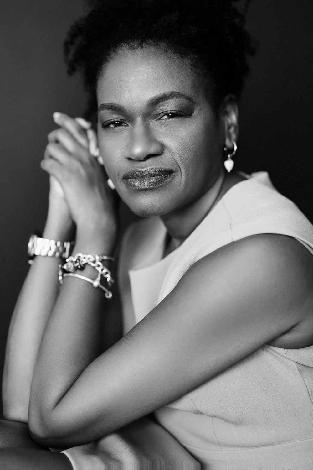 Black and white portrait of a woman with short, curly hair, wearing earrings, bracelets, and a sleeveless top, looking confidently at the camera.
