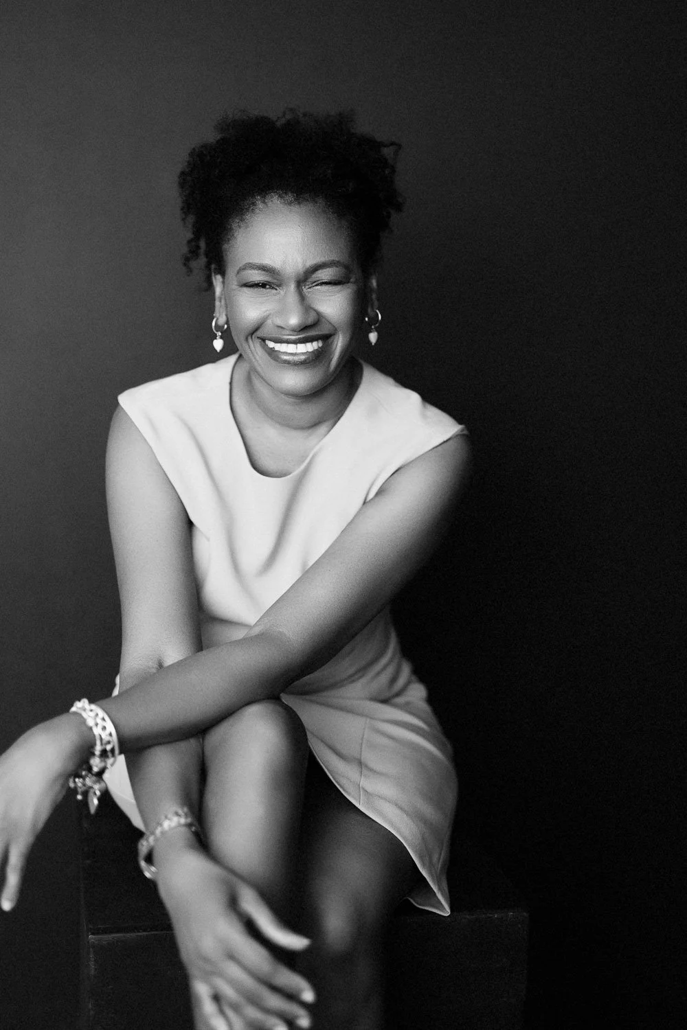 Black and white portrait of a smiling woman wearing a sleeveless dress, sitting with crossed legs, against a dark background. She has curly hair, earrings, and jewelry on her wrists.
