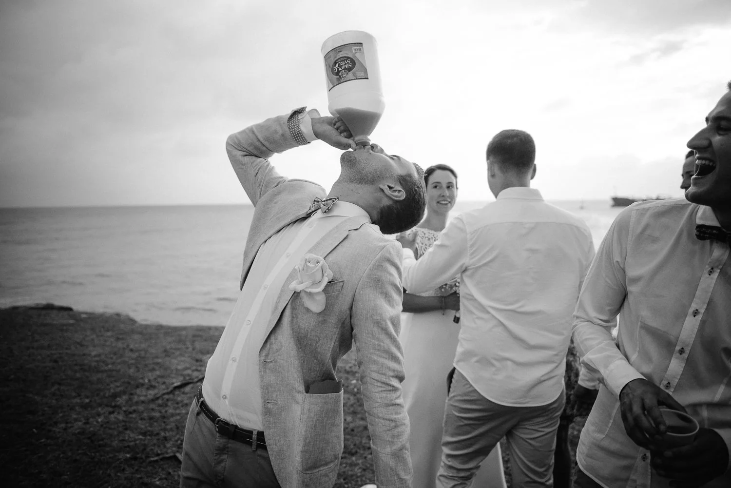 A man in a suit and bow tie is tilting his head back and drinking from a large bottle at an outdoor gathering near the water, with other happy people in formal attire around him.