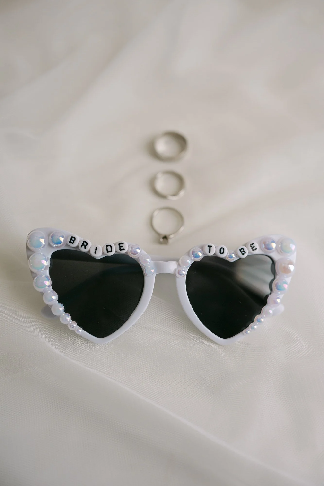 White heart-shaped sunglasses with beads spelling 'BRIDE TO BE' on the frame, placed on white fabric with three metal rings above.