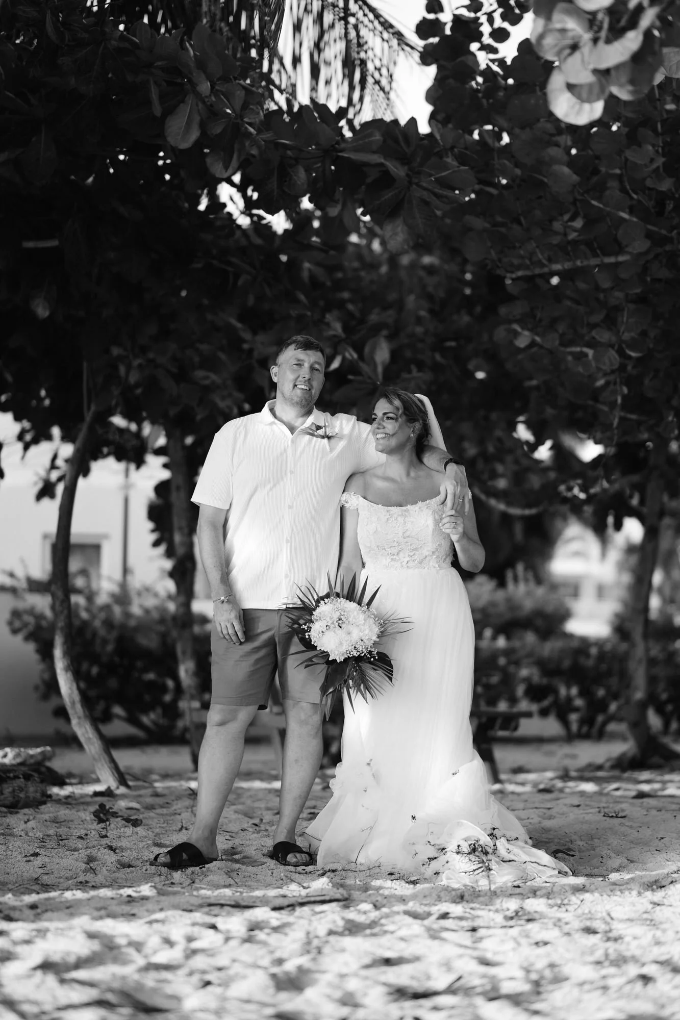 A black and white photo of a bride and groom standing together outdoors, smiling, with arms around each other. The bride is holding a bouquet of flowers, and they are standing on sandy ground beneath trees.