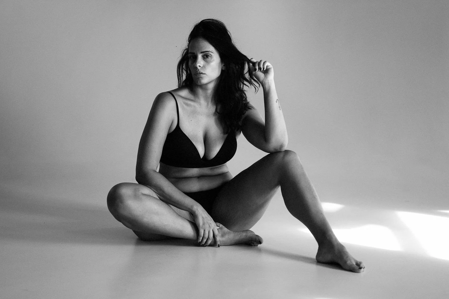 A black and white photo of a woman sitting on the floor, wearing a black bra and shorts. She has dark, wavy hair and is holding a lock of hair with one hand while looking into the camera.