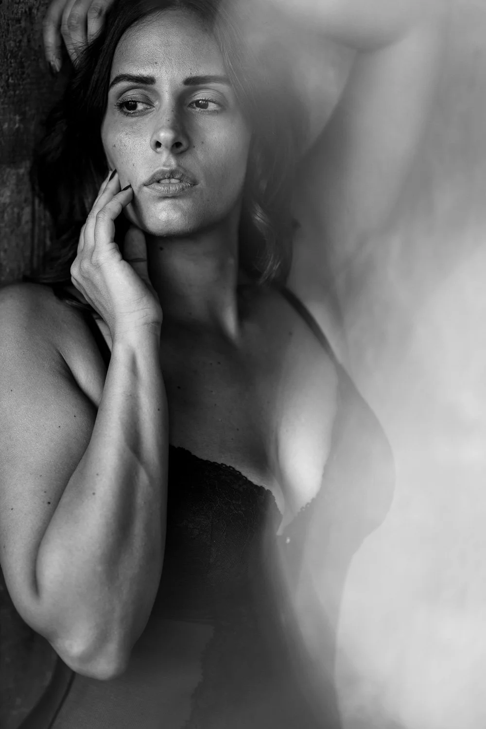 Black and white portrait of a woman with dark hair, touching her face with her left hand, wearing a black bra.