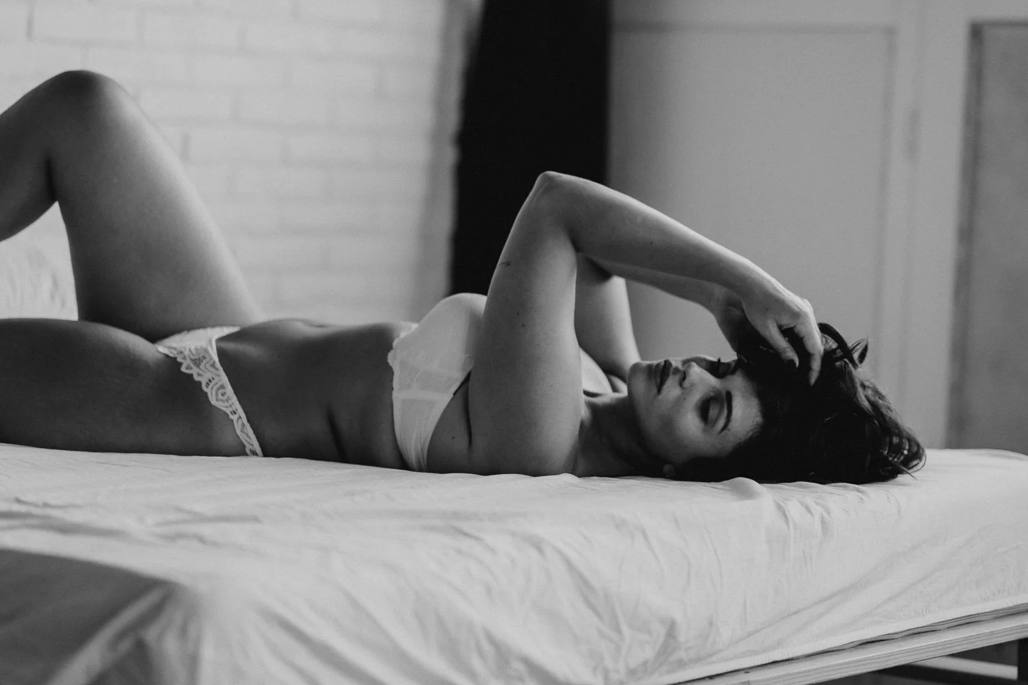 A woman lying on a bed, wearing lingerie, with one hand touching her hair and the other resting on her forehead, in a black-and-white photo.