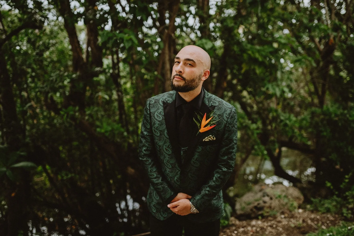 A man with a bald head and beard wearing a black and green paisley patterned suit jacket with a black shirt and tie, standing outdoors in front of green trees with water in the background, holding his hands in front of him with a flower boutonniere o