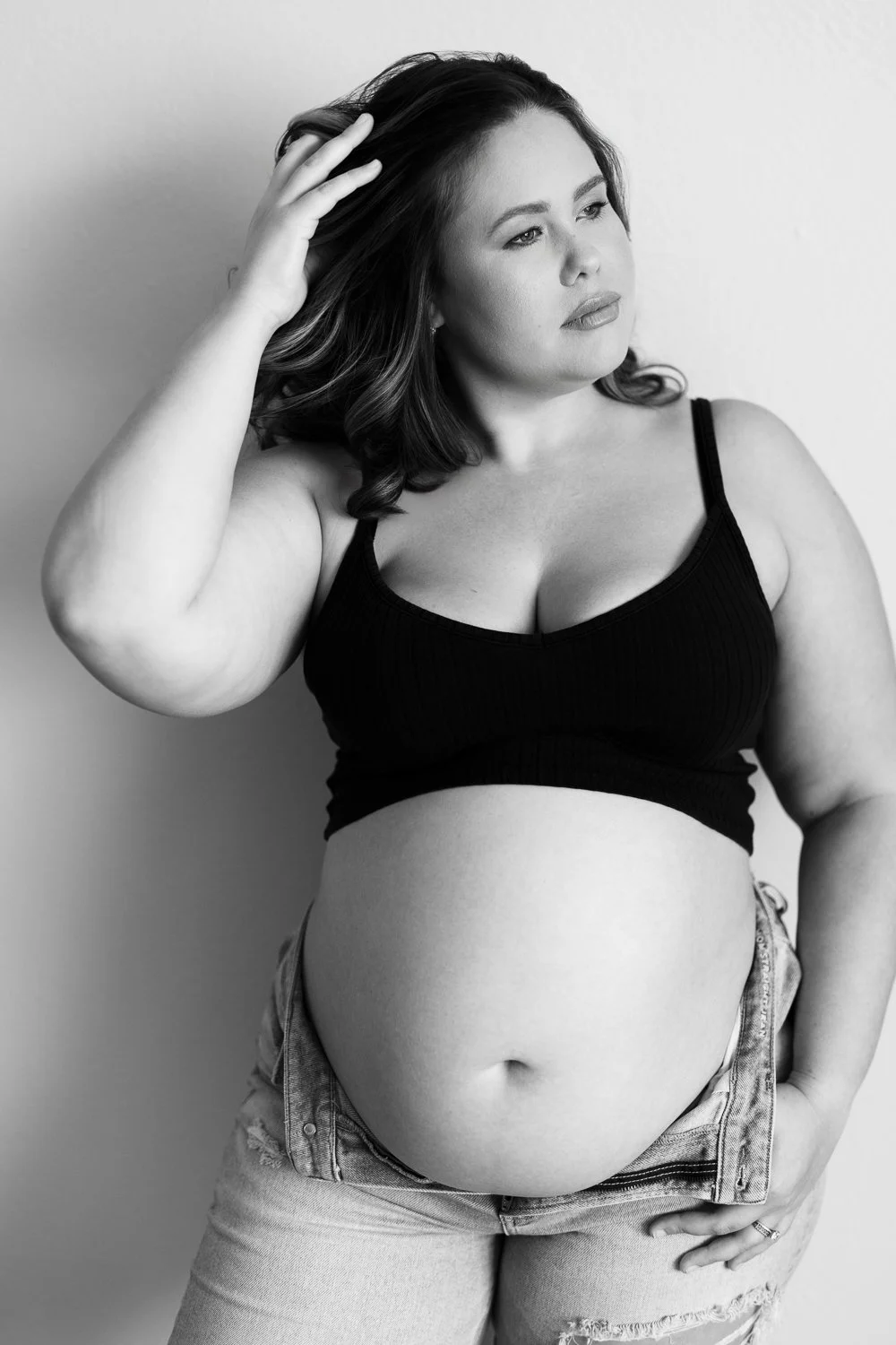 Black and white portrait of a plus-size woman with shoulder-length hair, wearing a black sleeveless top and distressed jeans, posing with her hand on her hair and looking thoughtfully to the side.