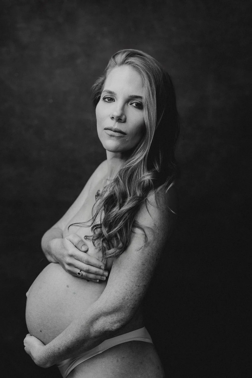 Black and white portrait of a pregnant woman with long, wavy hair, looking at the camera with a serious expression, against a dark background.