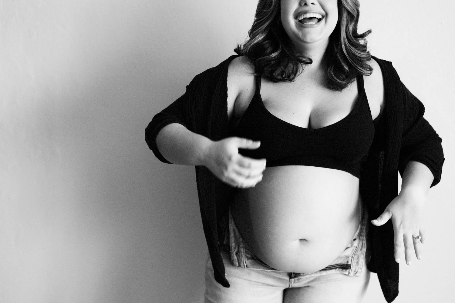 Black and white photo of a pregnant woman smiling, wearing a black sports bra and a black cardigan, with her hand resting on her pregnant belly.