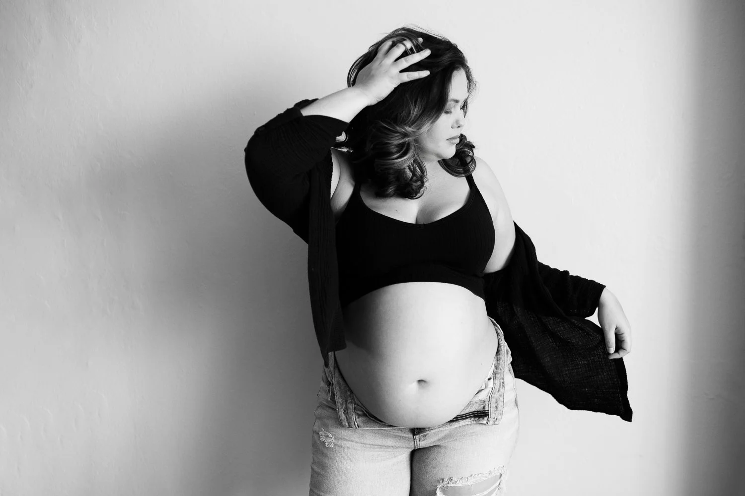 A pregnant woman standing against a plain wall, wearing a black crop top, ripped jeans, and a loose black cardigan, with her right hand on her head and her eyes closed.