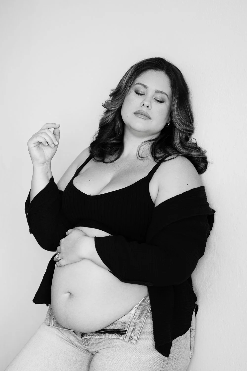 Black and white portrait of a woman with long dark wavy hair, wearing a black sleeveless top, a black jacket draped off her shoulders, and light-colored jeans, standing against a plain white wall.