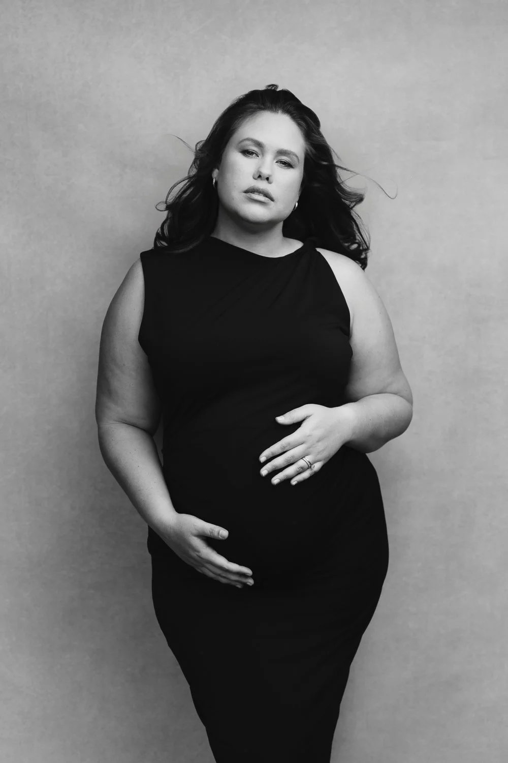 Black and white portrait of a pregnant woman in a sleeveless black dress, standing against a neutral background, with her hand resting on her belly.