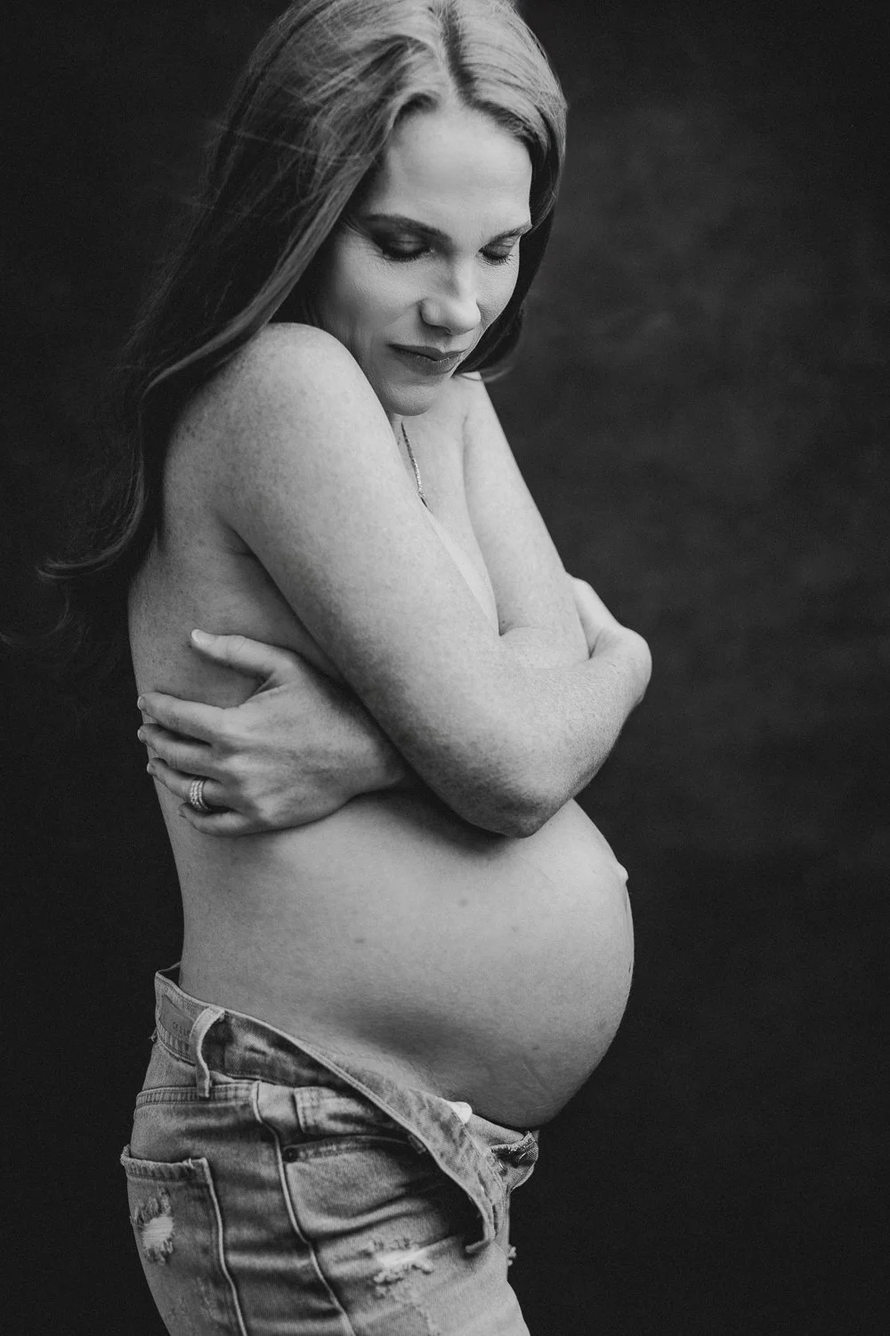 Black and white photograph of a pregnant woman with long hair, topless, wearing ripped jeans, and gently hugging her belly with her eyes closed.