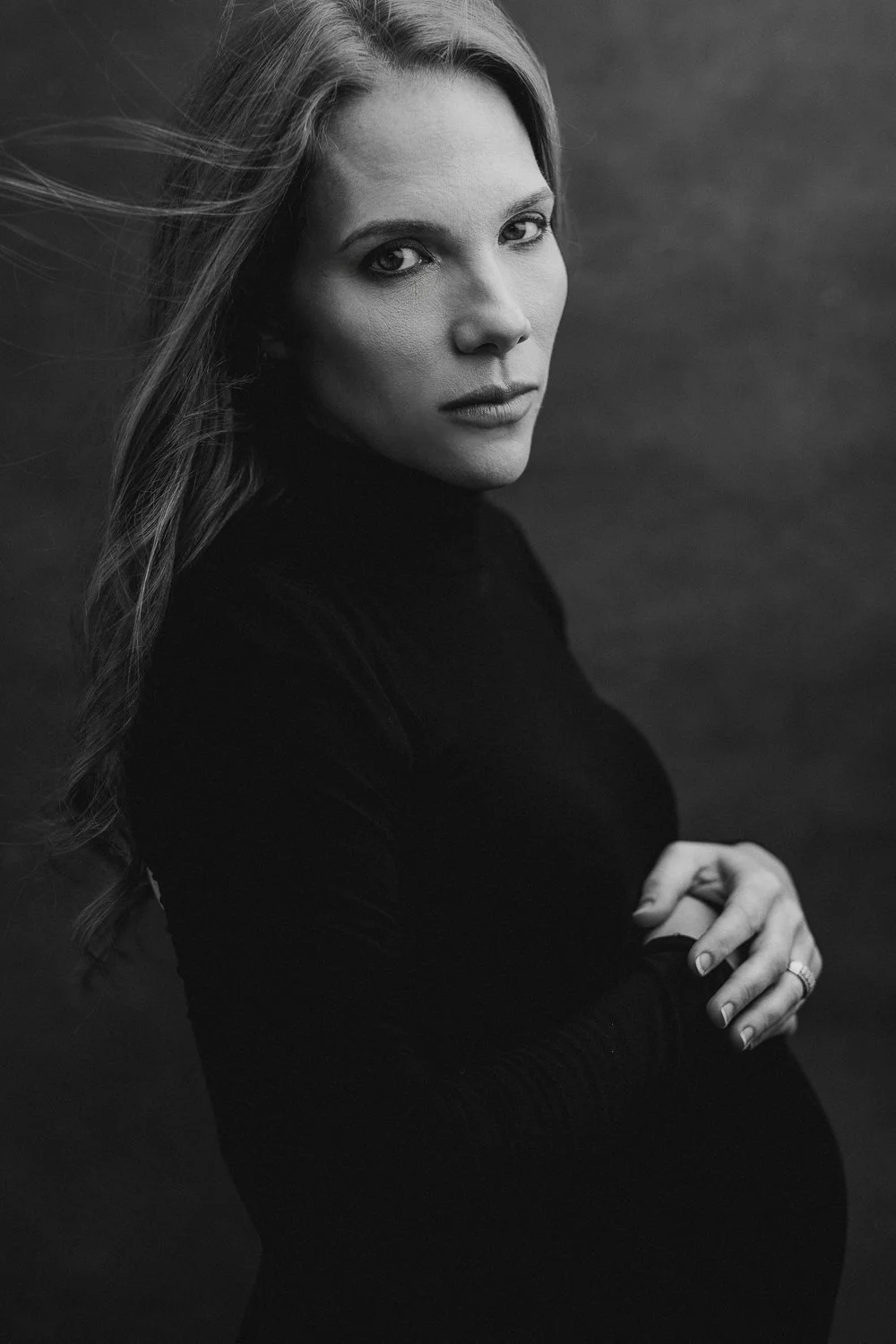 Black and white portrait of a pregnant woman with long, flowing hair, wearing a dark long-sleeve top, with her arms crossed over her belly, looking directly at the camera.