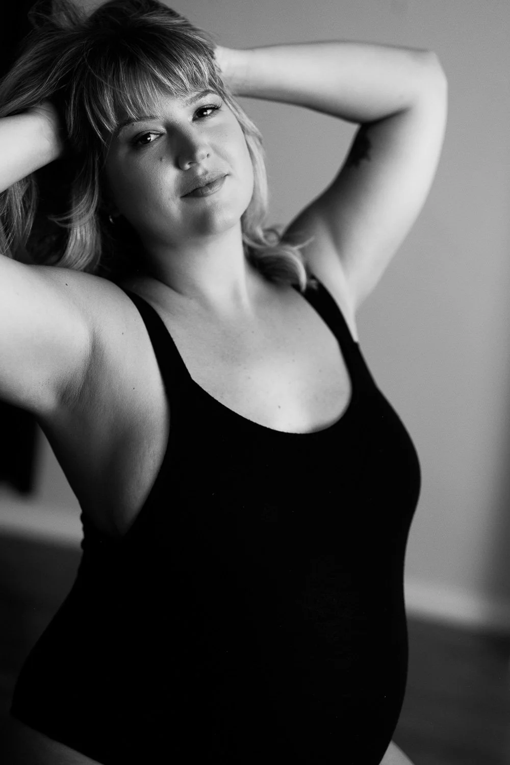 Black and white portrait of a woman with shoulder-length hair, wearing a sleeveless top, posing with her hands behind her head and smiling lightly.