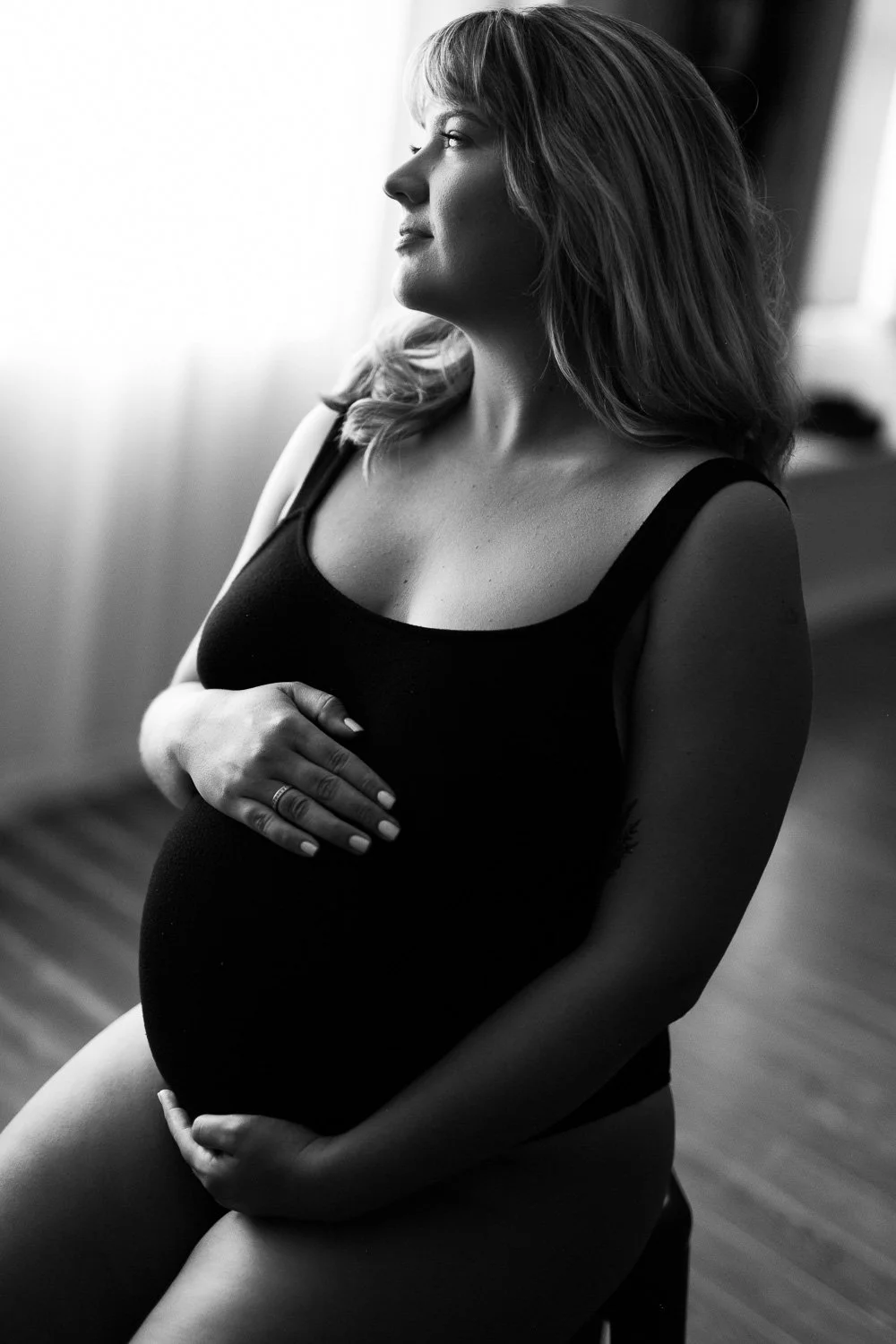 Black and white photo of a pregnant woman sitting and looking out a window, gently holding her belly.