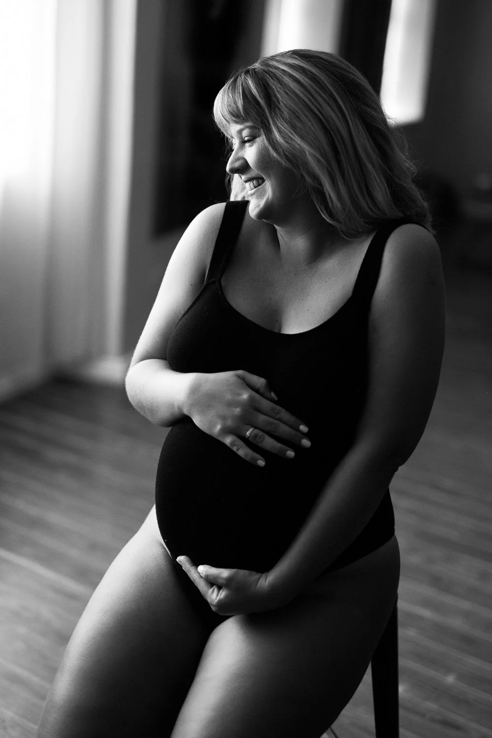 A pregnant woman smiling with her eyes closed, resting her hand on her belly, in a black and white photo indoors.