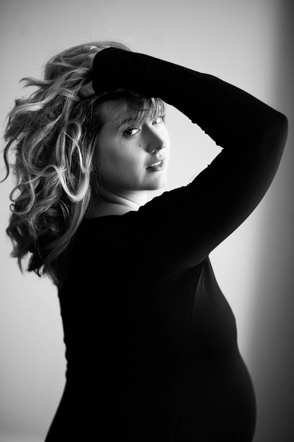 Black and white portrait of a woman with wavy hair, wearing a long-sleeved black top, posing with one arm raised and hand resting on her head, looking at the camera.