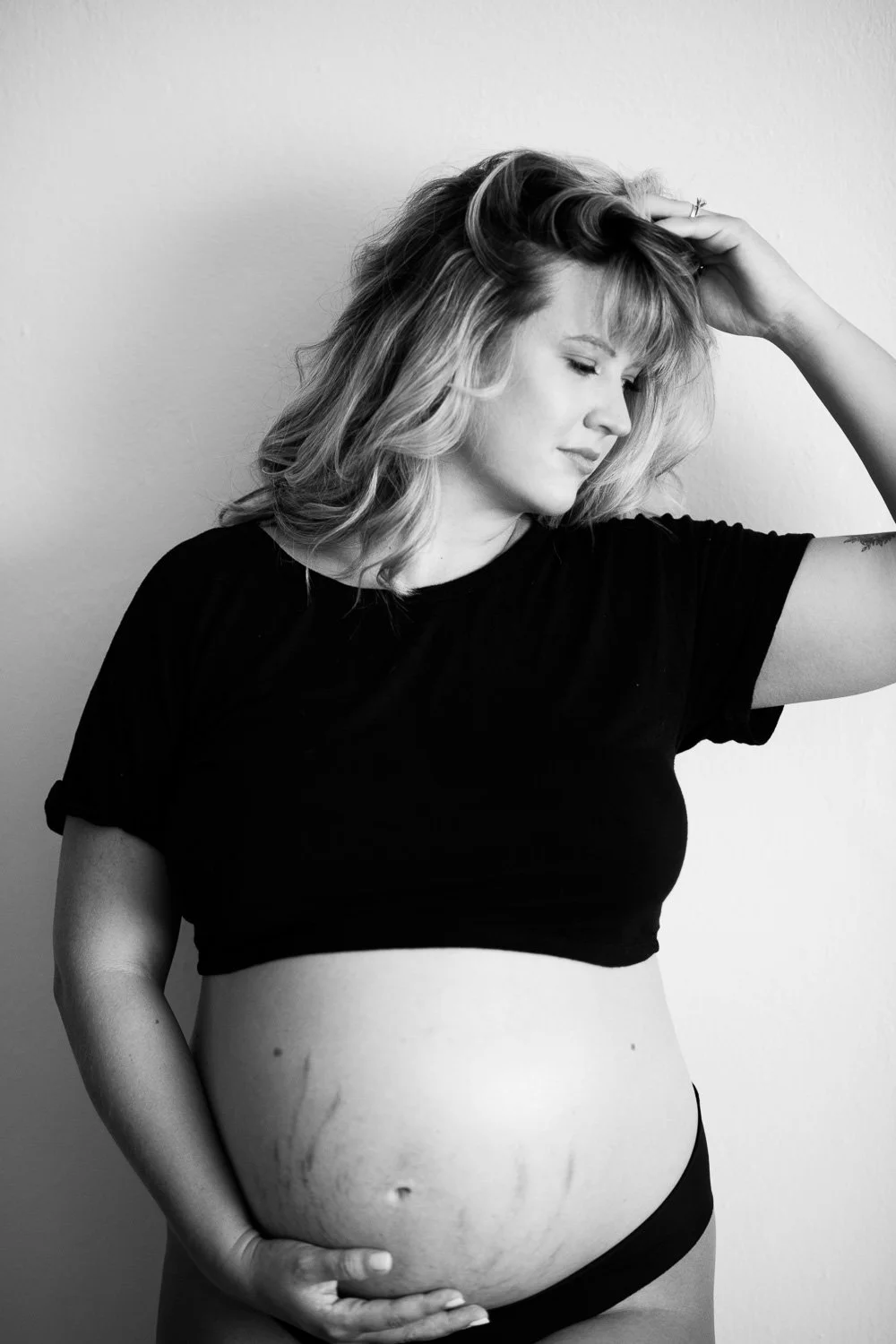 Black and white photo of a pregnant woman with curly hair, wearing a black t-shirt and underwear, standing against a plain background.