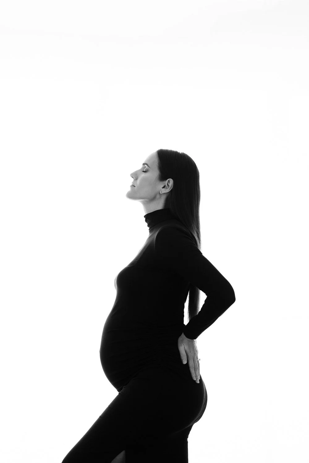 Silhouette of a pregnant woman in profile, wearing a black turtleneck and pants, with her eyes closed and one hand resting on her lower back against a plain, bright background.
