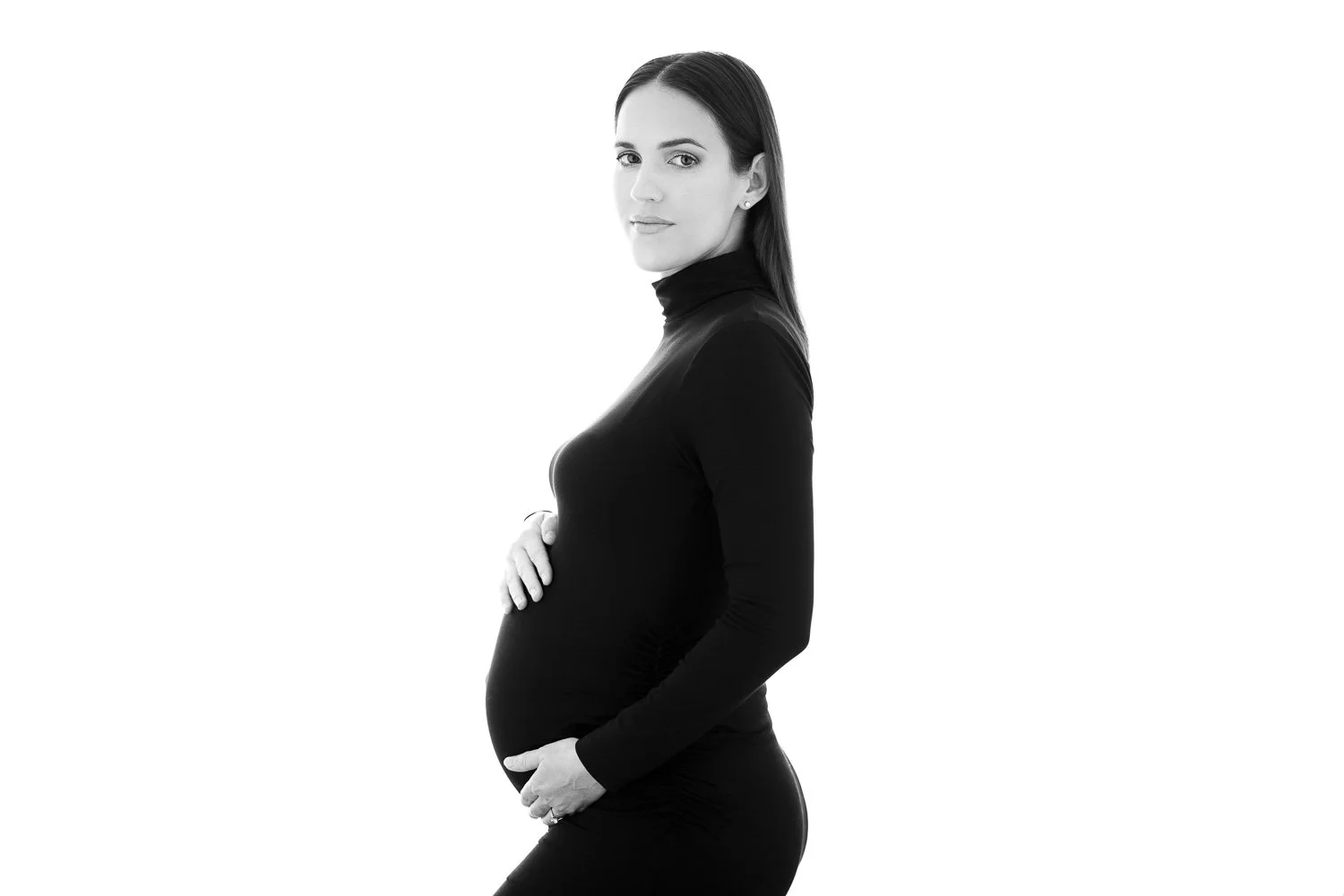 Black and white photo of a pregnant woman in a black long-sleeve turtleneck shirt and fitted pants, standing in profile with hands on her belly, looking at the camera.