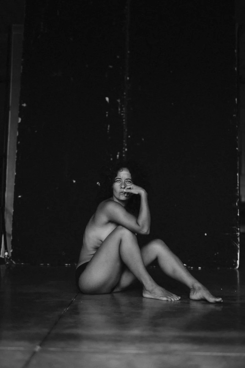 A black-and-white photo of a woman sitting on the floor against a dark wall, with one knee up and her hand near her face, looking contemplative or emotional.