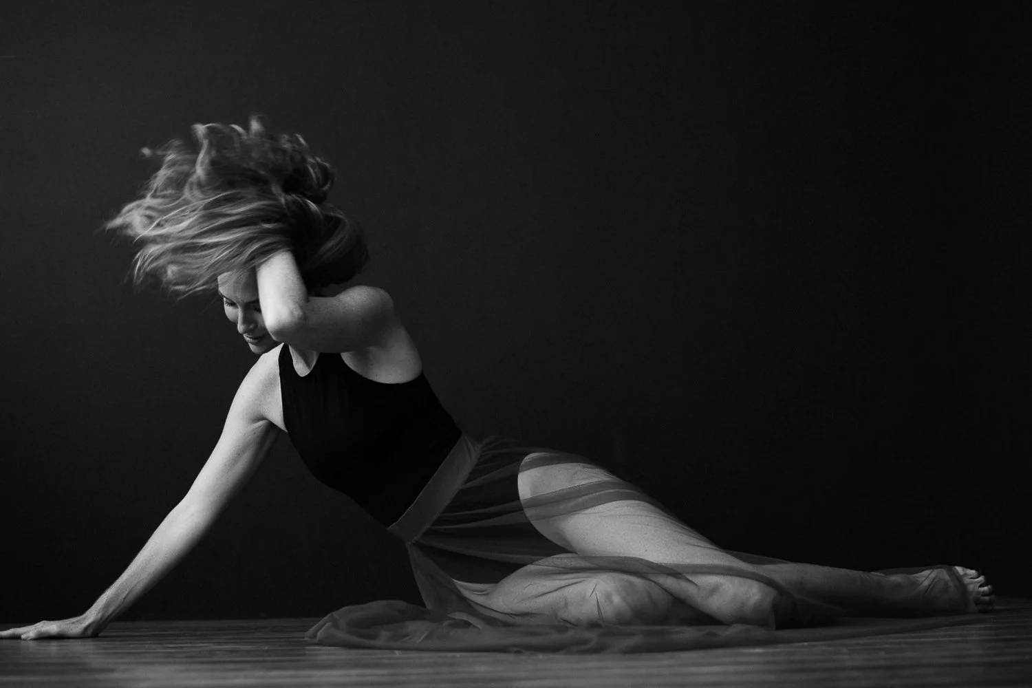 Black and white photo of a woman in a black top and transparent skirt, posing on the floor with her legs extended and her head tilted forward, hair flowing as she touches her head with her hand against a dark background.