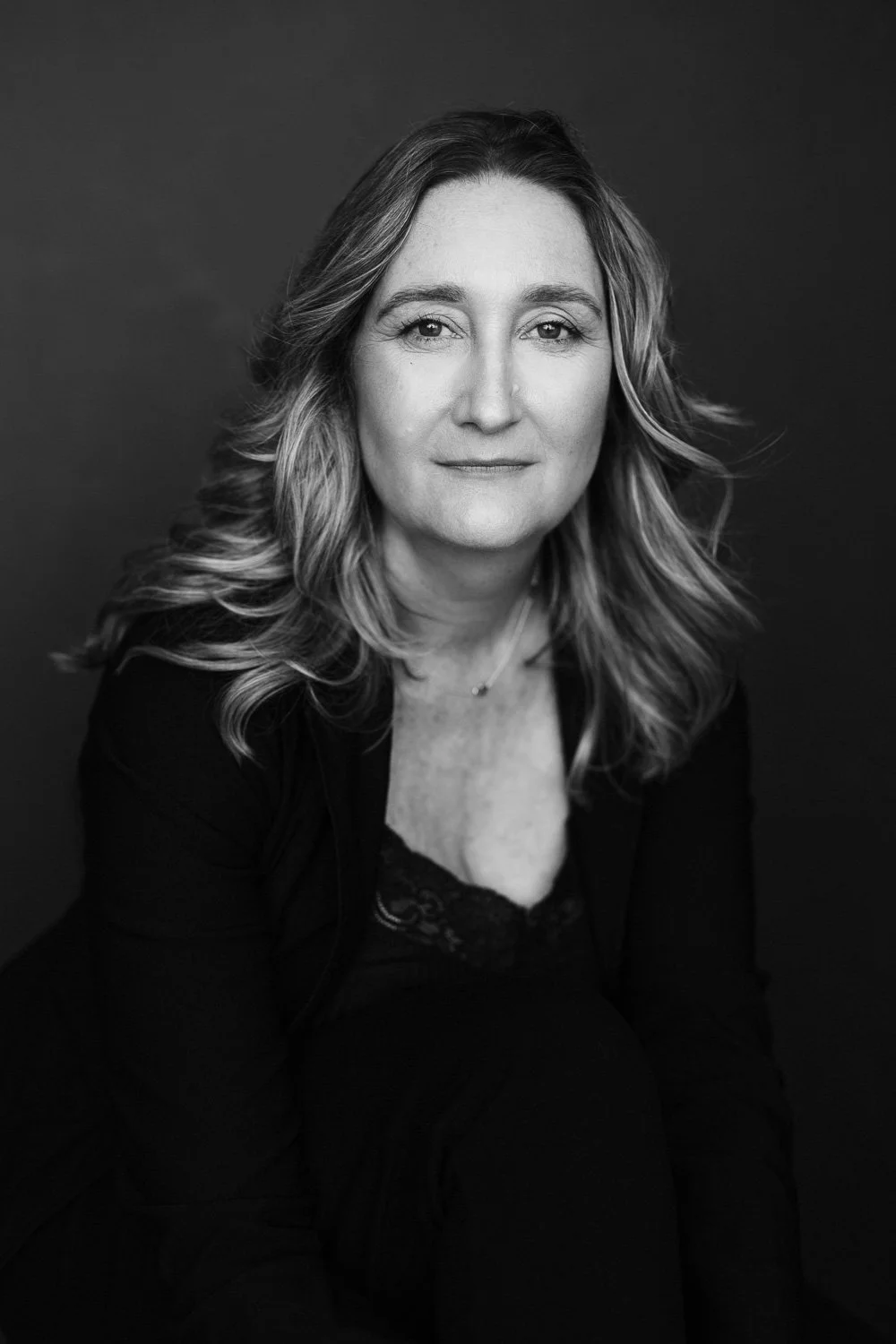 Black and white portrait of a woman with wavy hair, wearing a dark top with a lace trim, against a solid dark background.