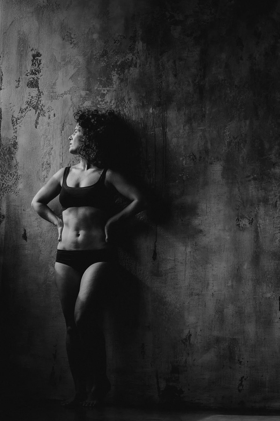 A woman in black underwear standing against a textured, dark wall with her hands on her hips, looking to the side.