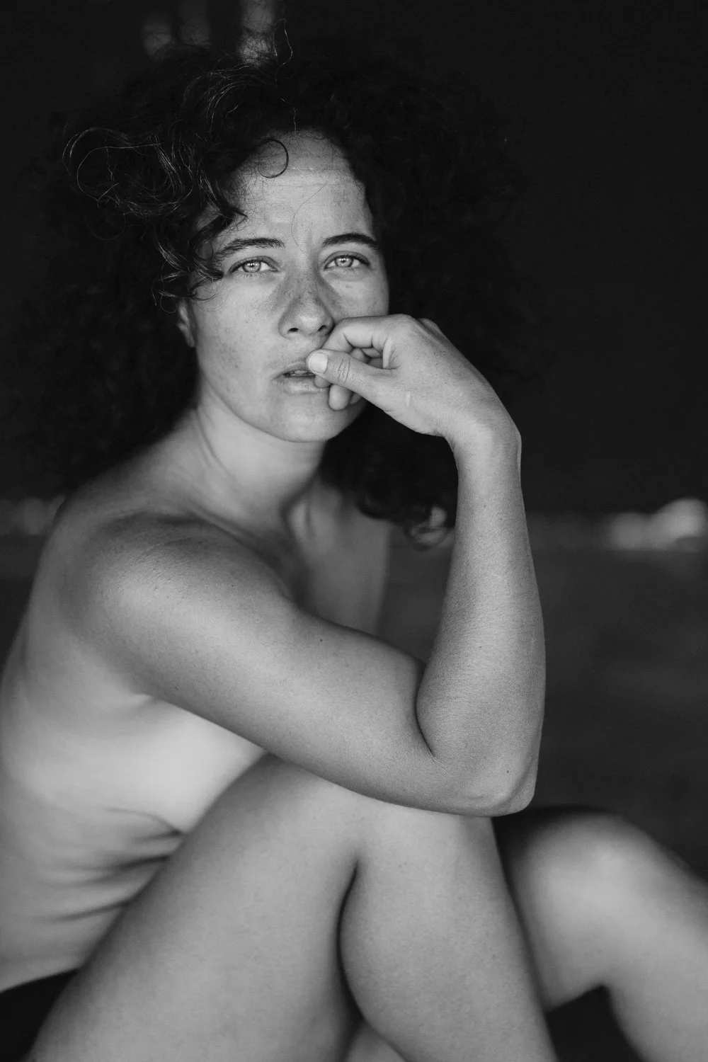 Black and white photo of a nude woman with curly hair, sitting with her knees drawn up, resting her chin on her hand, looking thoughtfully at the camera.