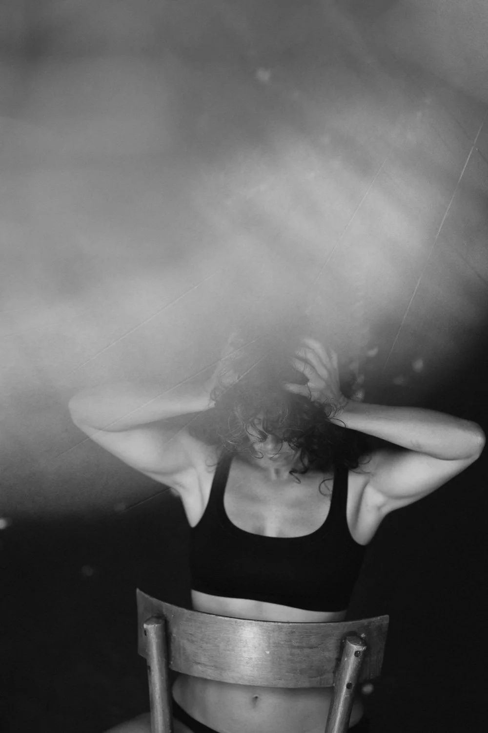 A woman in a black sports bra is sitting on a chair, with her hands in her hair, looking down, reflected in a mirror or water surface, in black and white