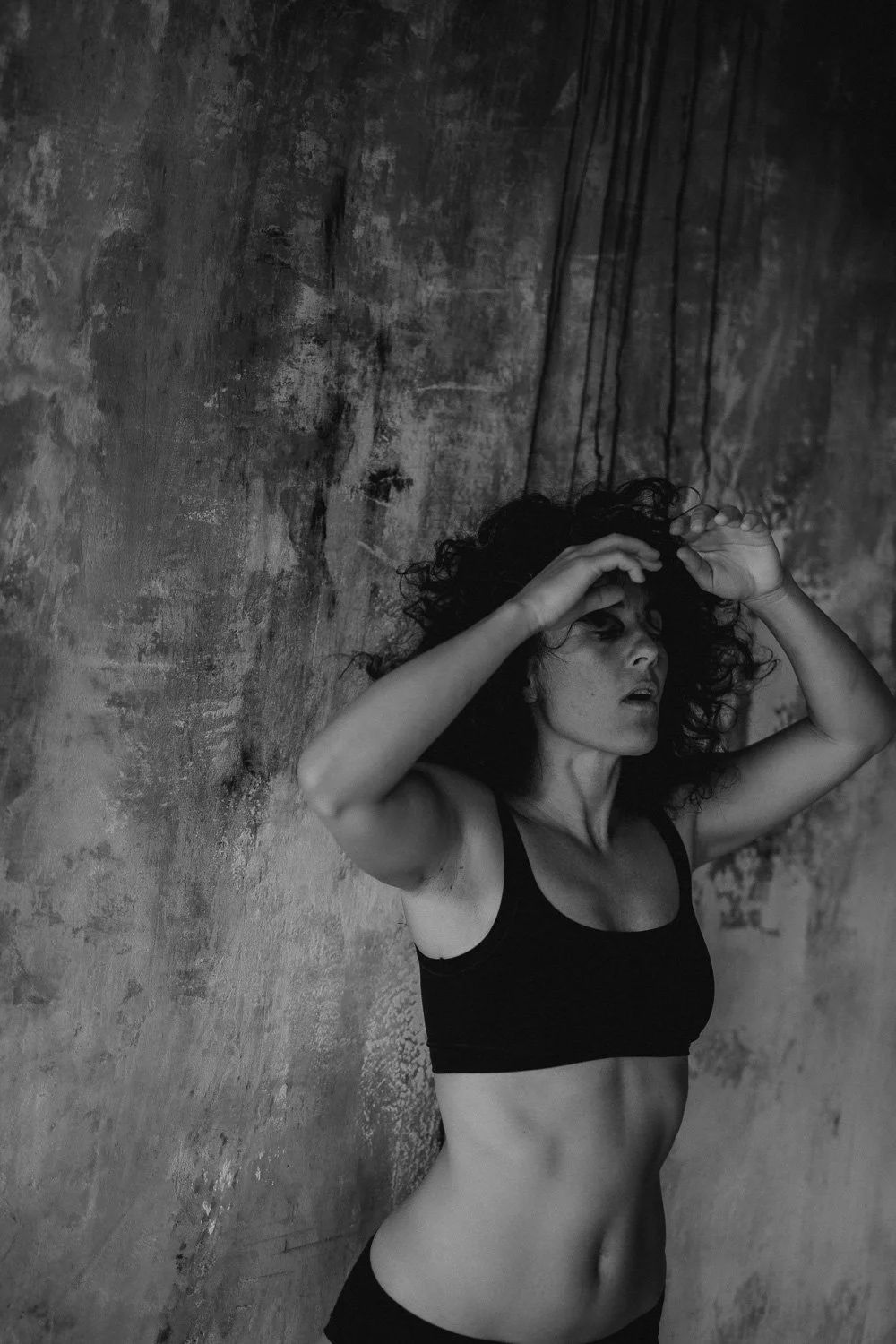Black and white photo of a woman with curly hair wearing a black sports bra, standing against a textured wall, with her hands on her forehead and a distressed expression.