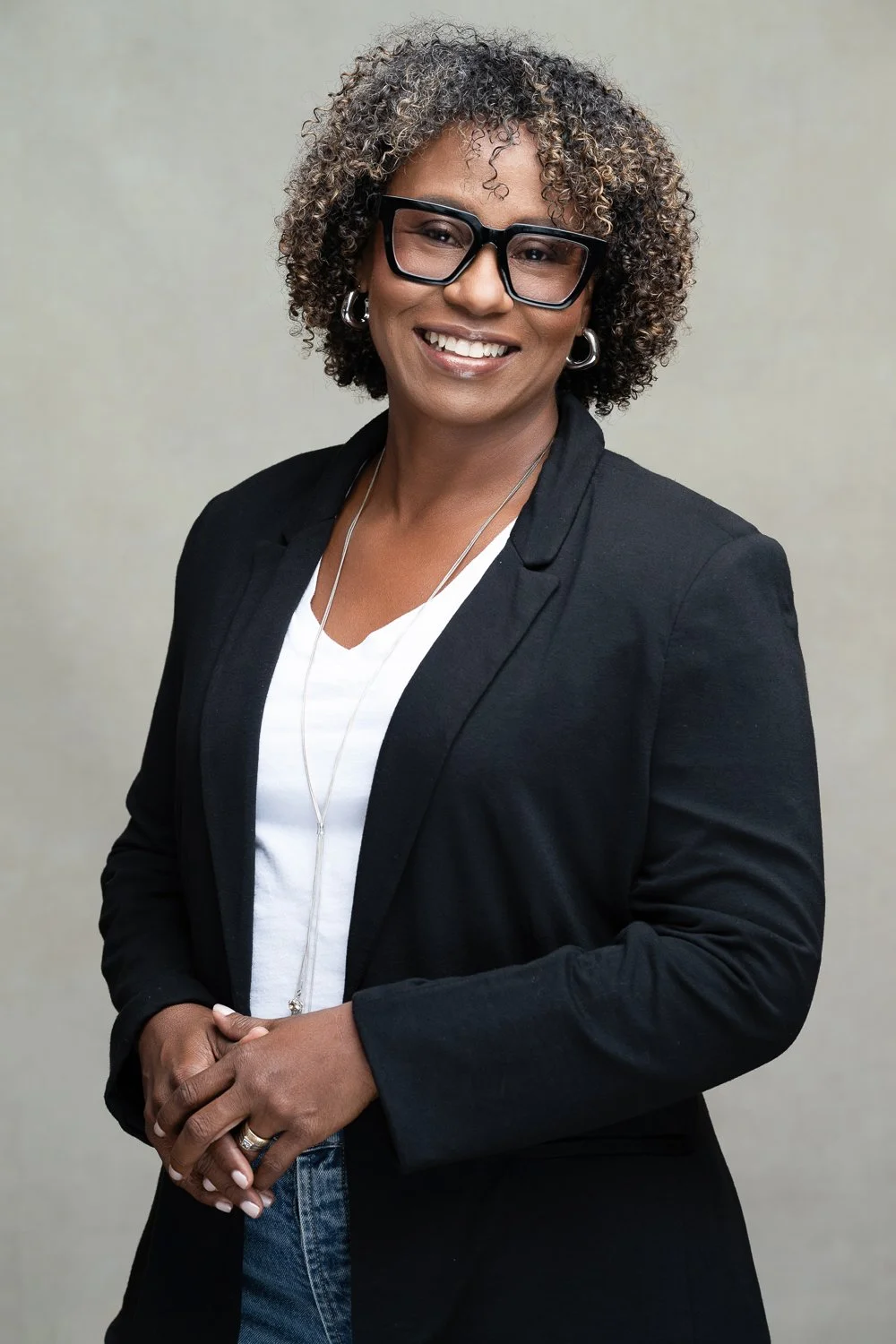 A woman with short curly hair, glasses, and hoop earrings, smiling and dressed in a black blazer over a white top, standing against a neutral background.