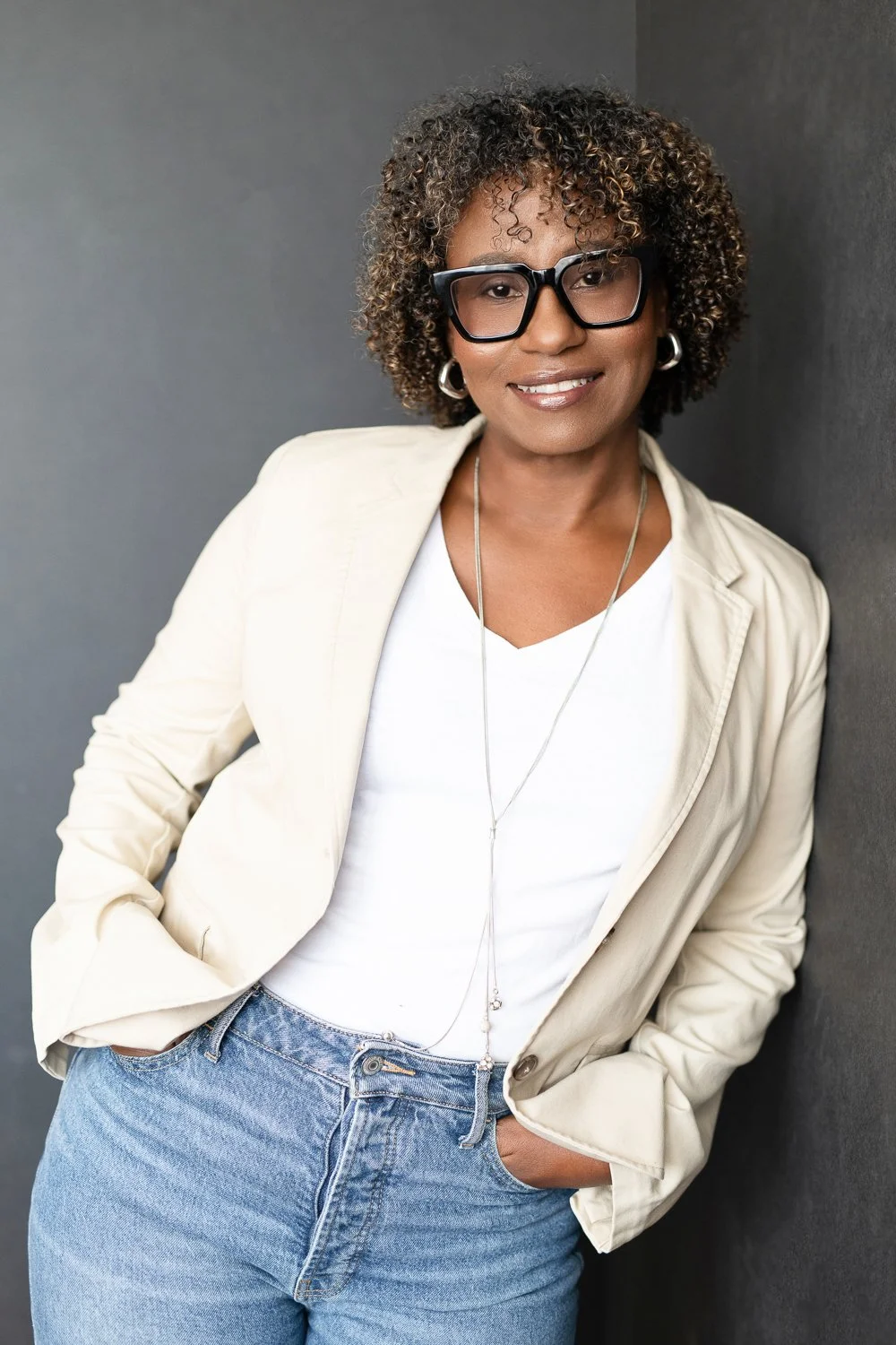 A woman with curly hair, glasses, and earrings smiling, wearing a white t-shirt, a light-colored blazer, and jeans, standing against a gray wall with her hands in her pockets.
