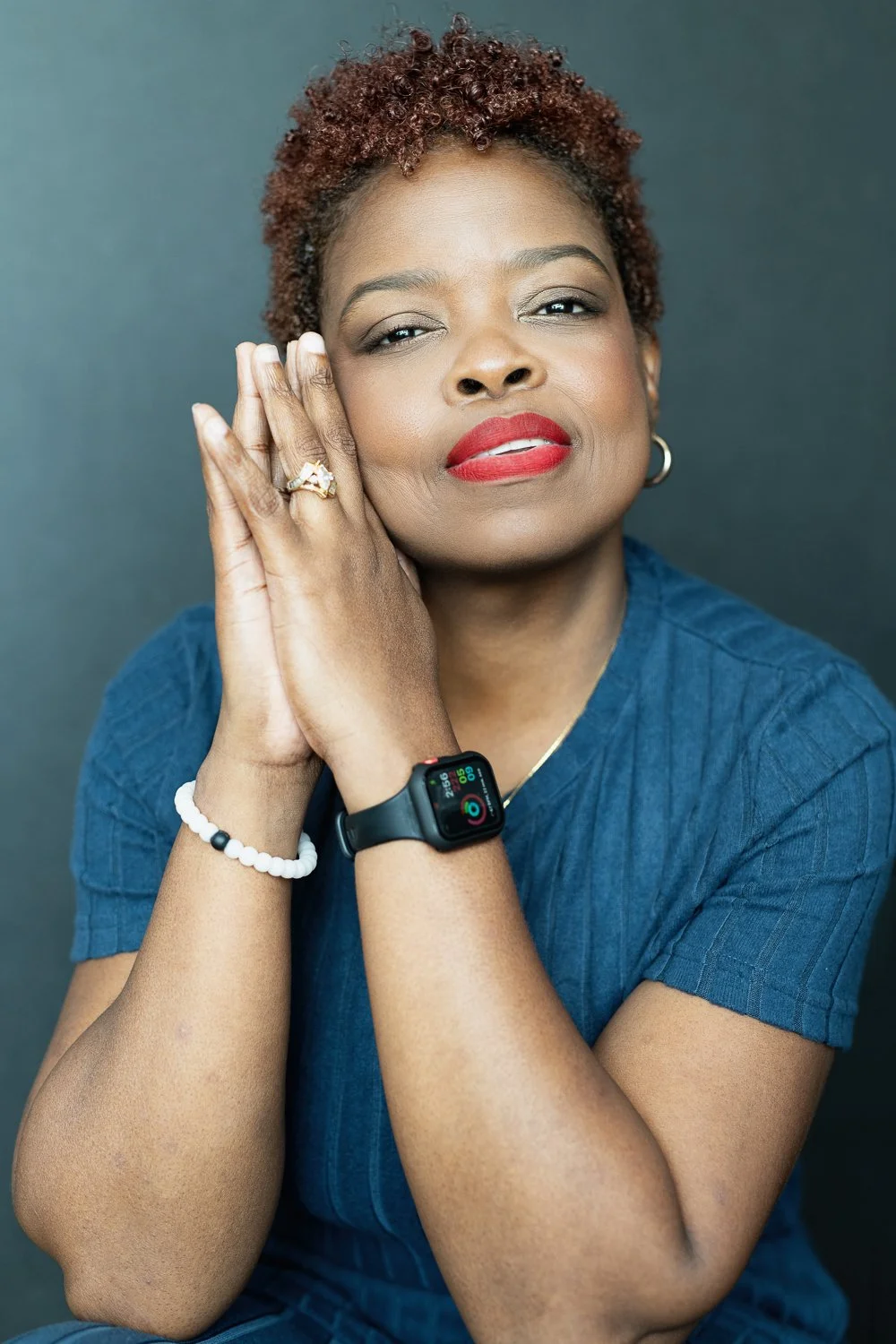 A woman with short curly hair wearing a blue shirt, red lipstick, and hoop earrings, resting her hand on the side of her face, showcasing a ring and bracelet, with a smartwatch on her wrist displaying health metrics.