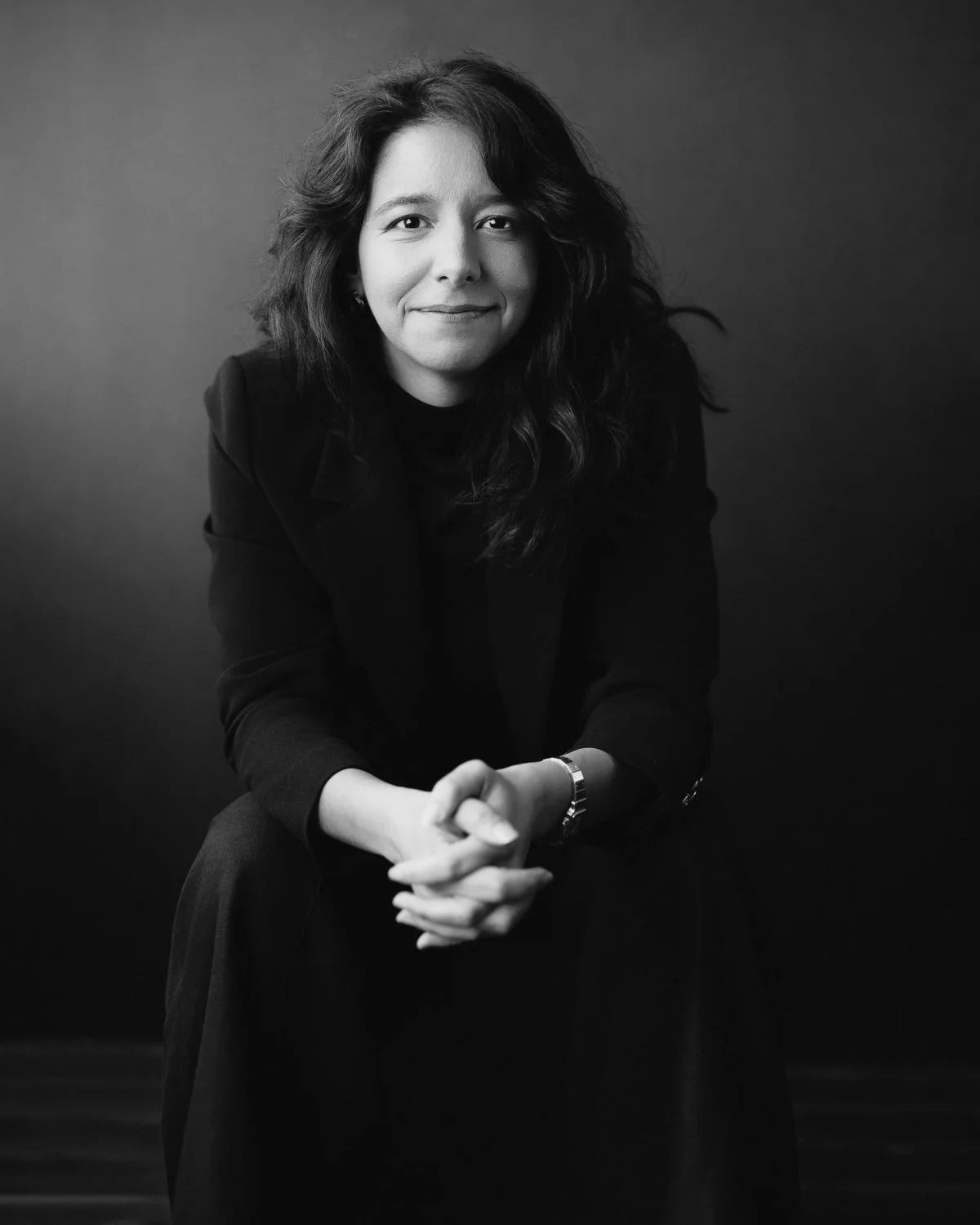 Black and white portrait of a woman with wavy hair, wearing a dark blazer, sitting with her hands clasped, smiling slightly, against a dark background.