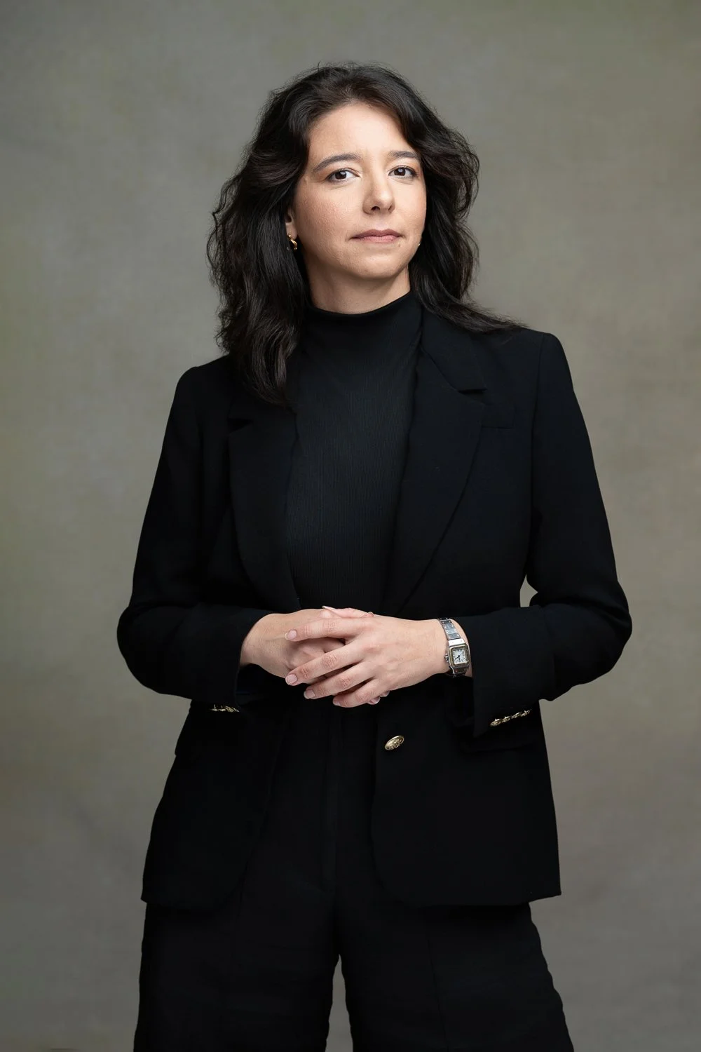 A woman with dark, wavy hair wearing a black turtleneck and blazer, standing against a neutral background, looking confidently at the camera.