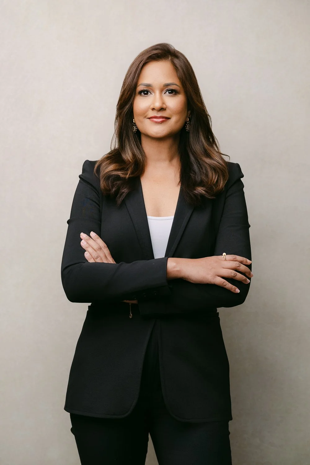A confident woman with shoulder-length brown hair, wearing a black blazer over a white top, standing with arms crossed against a plain beige background.