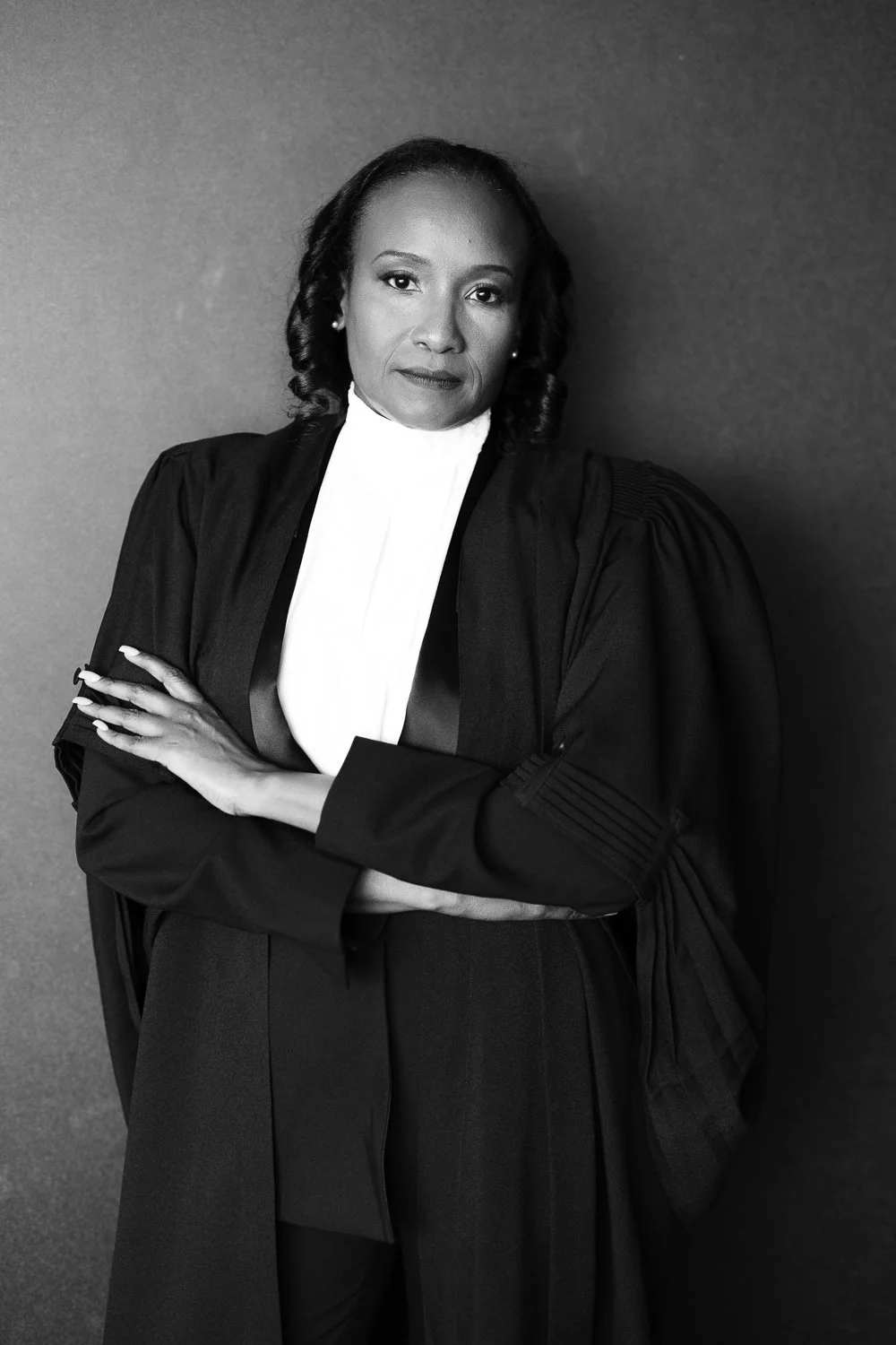Professional portrait of a woman in a courtroom lawyer's robe, crossing her arms and looking confidently at the camera, black and white photo.