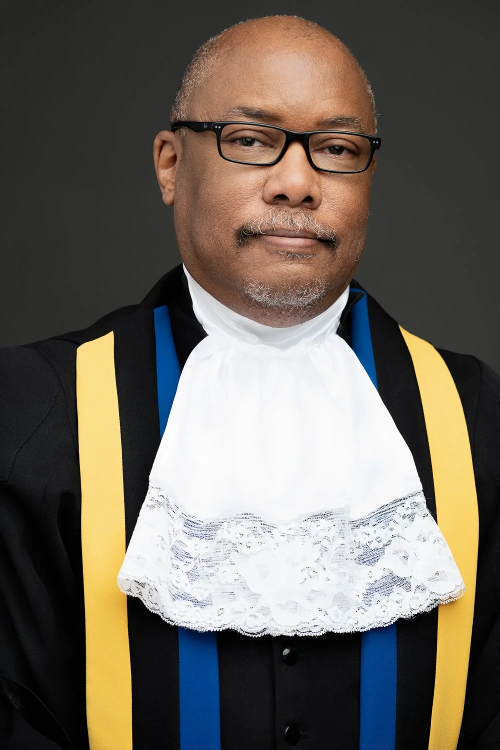 A judge wearing glasses, a black judicial robe with yellow and blue accents, and a white lace jabot, standing against a dark background.