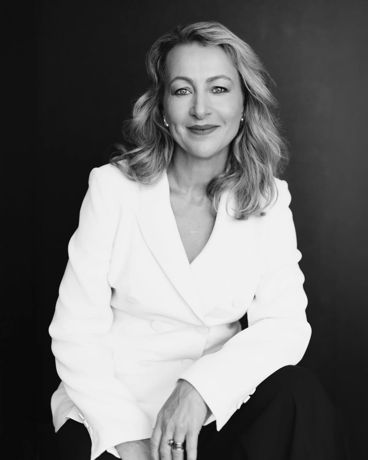 Black and white portrait of a woman with wavy hair, wearing a white blazer and black pants, sitting against a dark background.