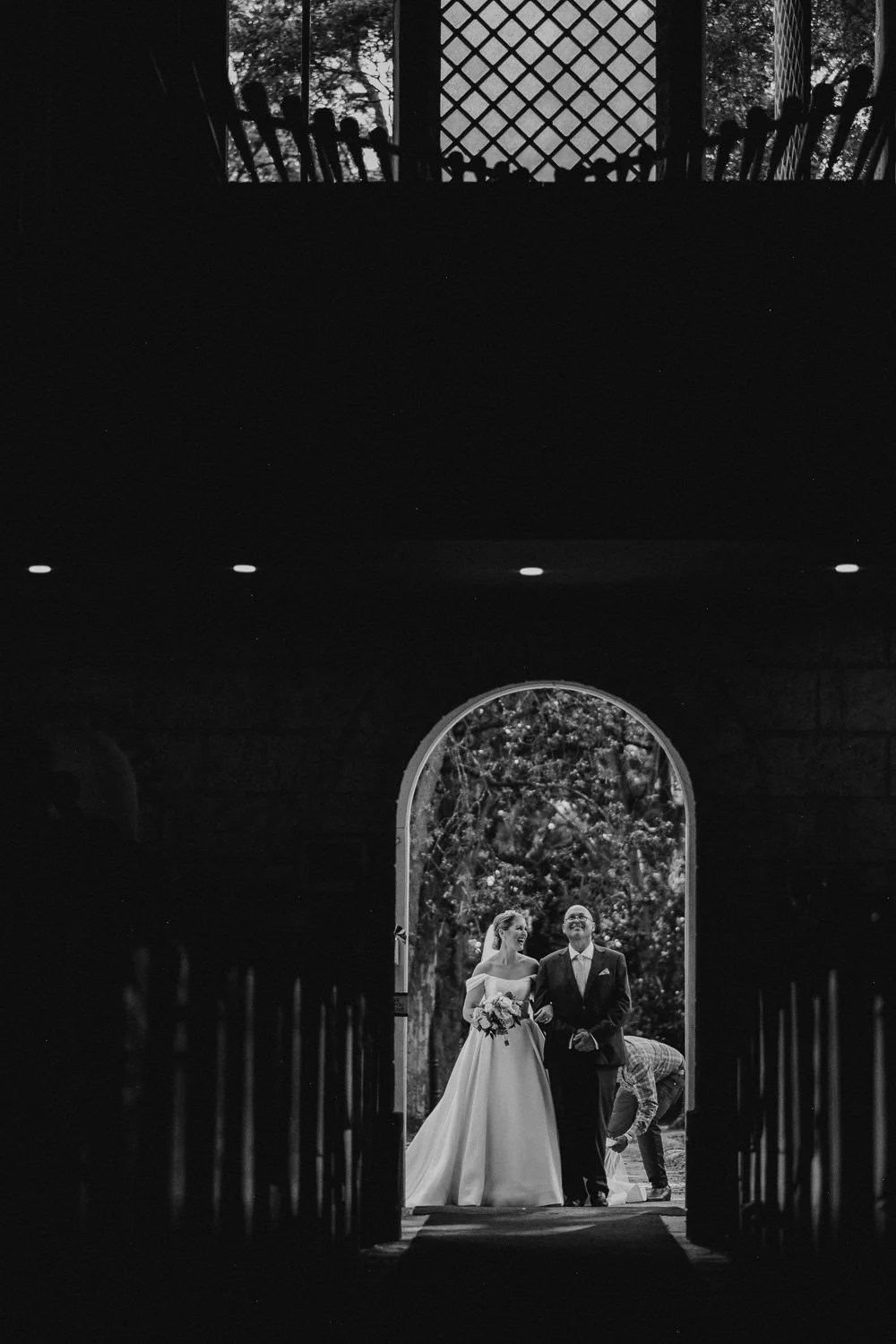 Black and white photo of a smiling bride and groom walking through a church arched doorway, holding hands, with a person adjusting the bride's dress in the background.