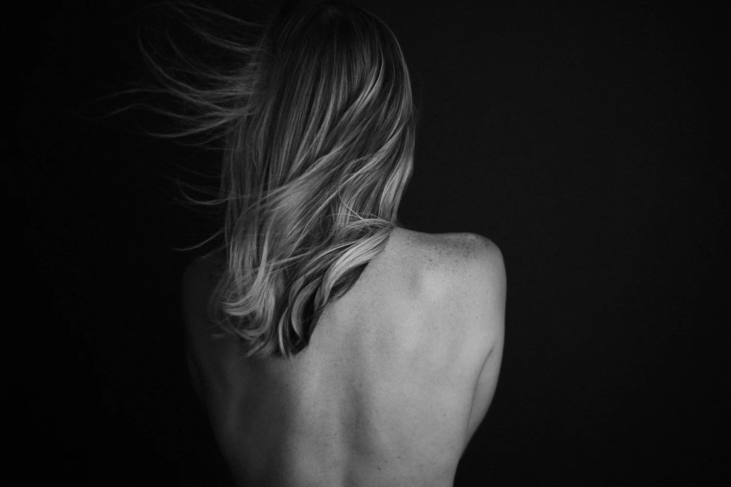 Black and white photo of a woman with long, wavy hair turned away from the camera, showing her back and shoulder against a dark background.