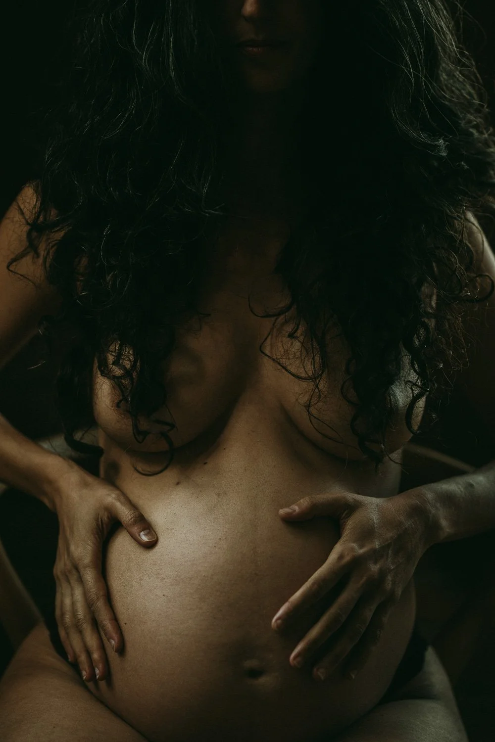 A person with long, dark curly hair, topless, holding their pregnant belly in a dimly lit environment.