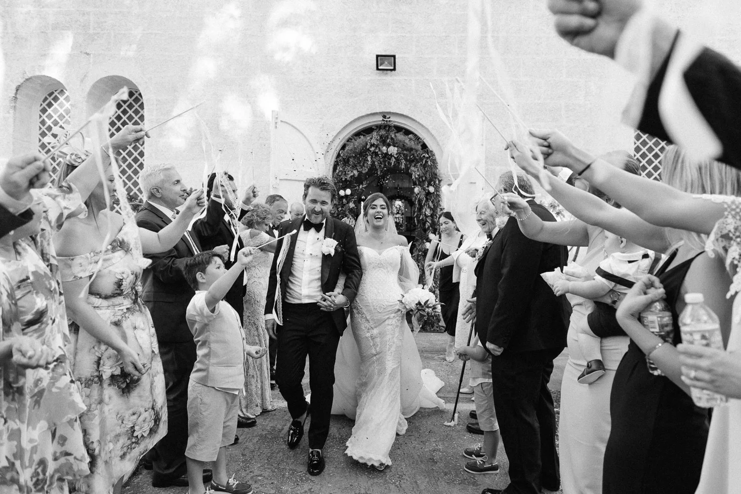 Black and white photo of a newlywed couple walking out of a church, smiling and holding hands, surrounded by guests celebrating with confetti and streamers.