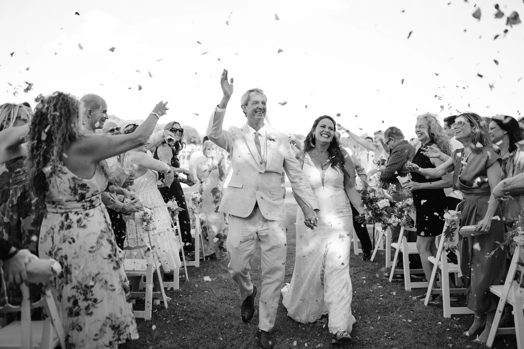 Black and white photo of a wedding celebration with a couple walking down an aisle, holding hands and smiling, surrounded by guests throwing confetti.