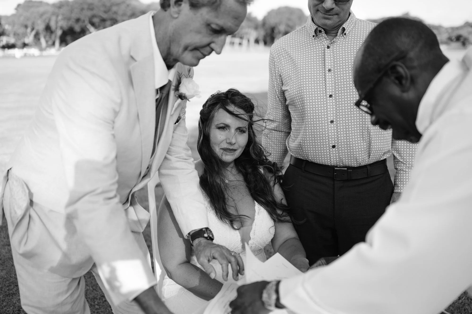 A black-and-white photo of a woman in a wedding dress holding papers, surrounded by four men, including one in a white suit with a flower on the lapel, outside in a park or open field.